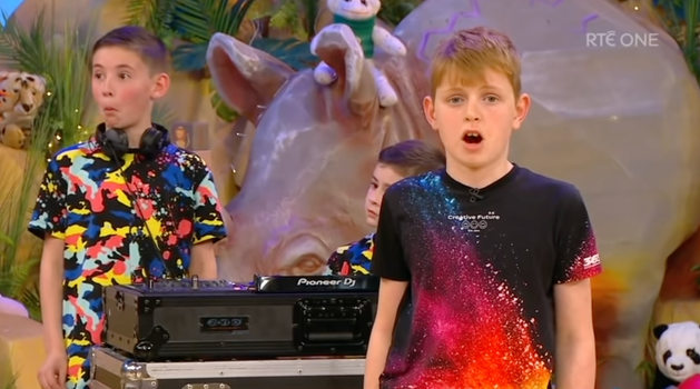 Two little boys behind a set of DJ decks on the toy show set, with one boy in front singing