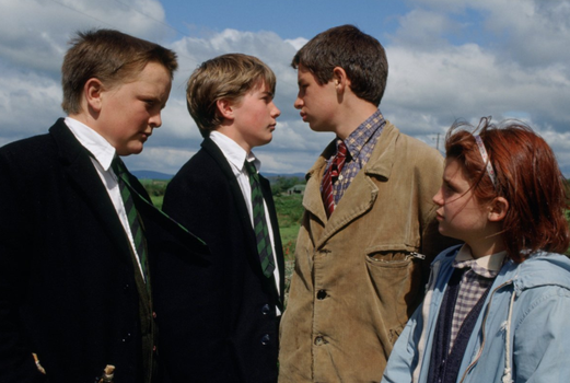 Still from War of the Buttons. Two boys in school uniforms face a taller boy in a brown jacket accompanied by a little girl with red hair.