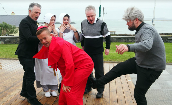 Man dressed as Bishop Brennan from Father Ted (red cloak and cap) being kicked up the arse by a man dressed as Father Ted (grey wig, grey jumper etc)