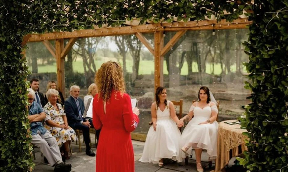 scene from an outdoor wedding with greenery and wooden beams in the background - two brides holding hands, and an officiator reading from a book