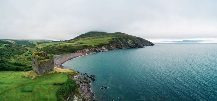 aerial shot of Dingle Peninsula and Minard Castle, with blue seas, grassy cliffs, a rocky beach and cloudy sky