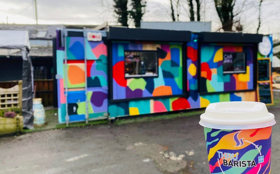 converted shipping container painted with multicoloured shapes, a thru barista cup with similar design is held in the forgeground