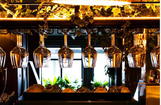 wine glasses hanging upside down on a rack in a restaurant, coffee machine underneath and window with sun shining through in the background
