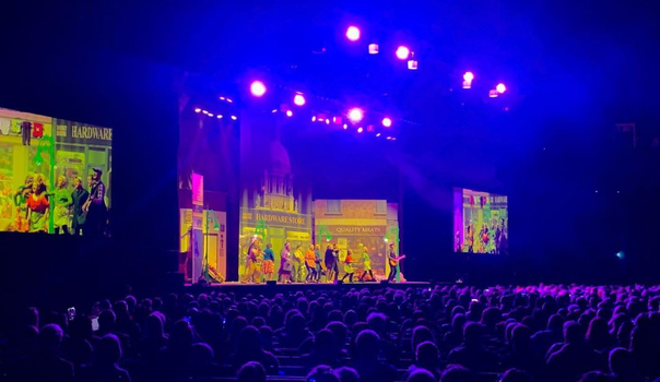 busy stadium with Mrs Browns Boy cast on stage and large monitors on either side of them
