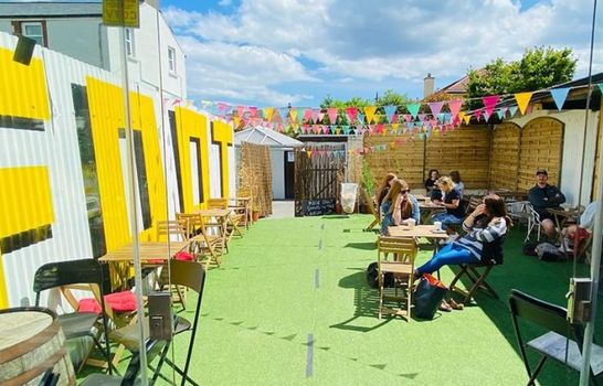 outdoor dining area with artificial grass, colourful flags and tables and chairs. Blue skies in the background