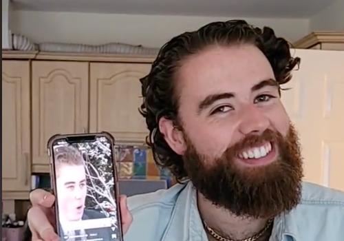 man smiling for camera holding a phone showing a younger version of him being interviewed in the snow