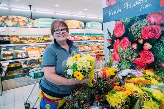 older woman with glasses smiling and arranging flowers at display in lidl