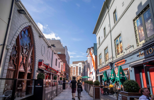 two people walking down a street in Cork City with bars and restaurants with outdoor seating and umbrellas on either side of them