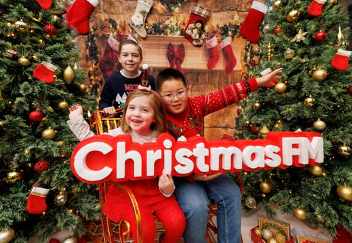 children in front of a fireplace decorated for christmas and christmas trees on either side, holding a sign that says Christmas FM