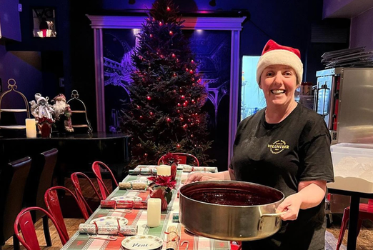 a woman holding a large pot of food with christmas tree and table behind her