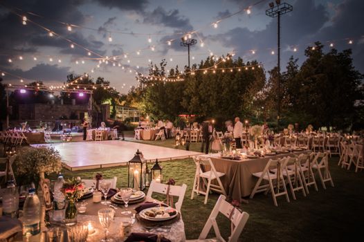 outdoor wedding scene in a field surrounded by trees, a dancefloor with tables and chairs and fairylights overhead